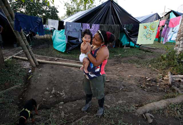 A woman holds her smiling baby, in the background are clothes strung along a clothesline and a tent.