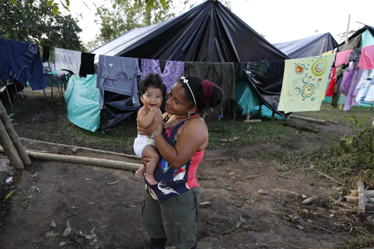 A woman holds her smiling baby, in the background are clothes strung along a clothesline and a tent.