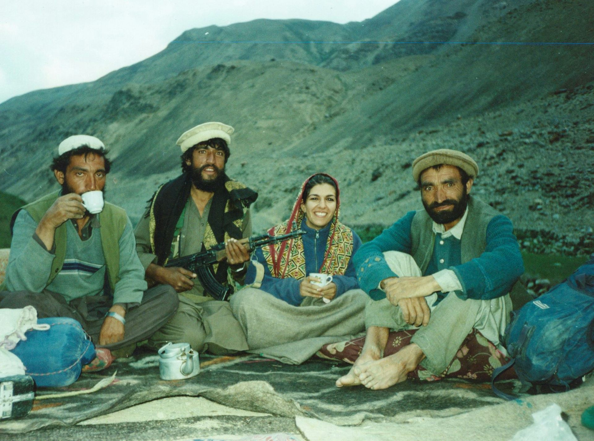 A woman in a headscarf and three men in turbans sit on the floor, smiling with a mountain in the background.