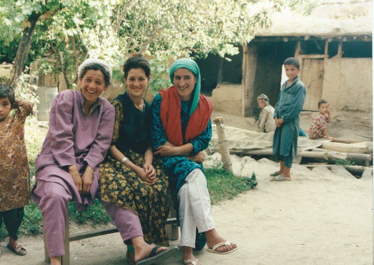 Three laughing women sit on a bench by a tree with children in the background.