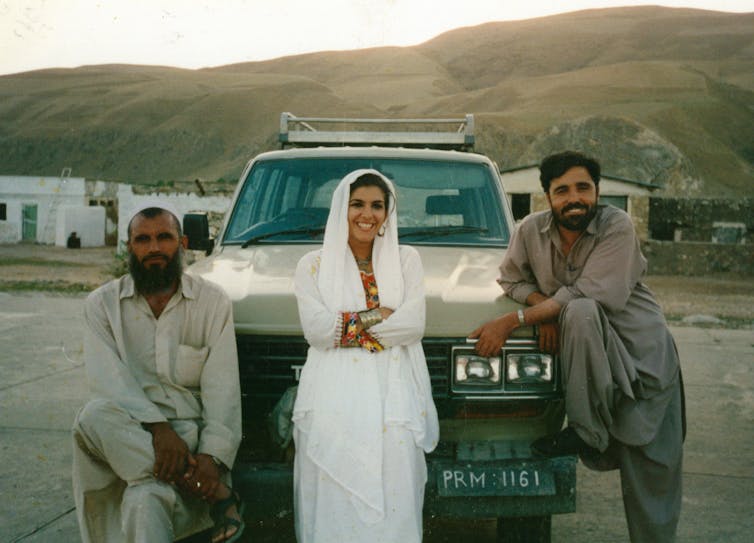 A woman wearing white leans against a car with two men.
