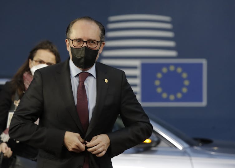 Alexander Schallenberg poartă o mască de față la un eveniment european.  Un steag al UE este ilustrat pe peretele din spatele lui.