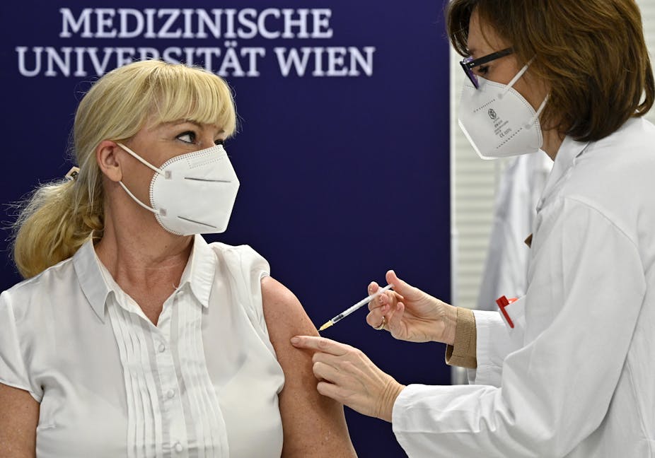 A female medical worker gives a vaccine to another female medical worker. The wall behind them says they are at Vienna's Medical University.