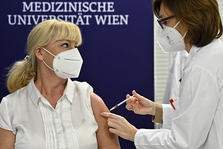 A female medical worker gives a vaccine to another female medical worker. The wall behind them says they are at Vienna's Medical University.