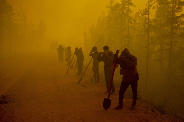 A row of people holding shovels stand on the edge of a dirt road, surrounded by orange smoke.