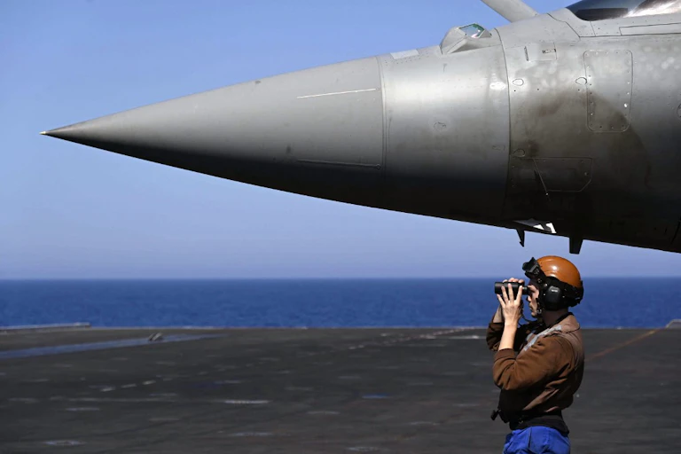 Un militaire sur le pont d'un porte-avions regarde vers la gauche à la jumelle en dessous du nez d'un Rafale.
