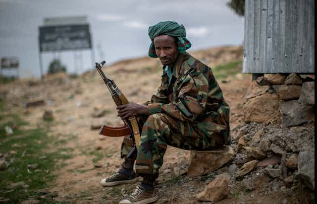 A man in army fatigues sits on a rock while holding a gun.