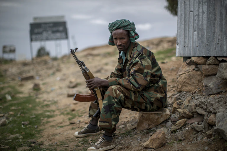 A man in army fatigues sits on a rock while holding a gun.