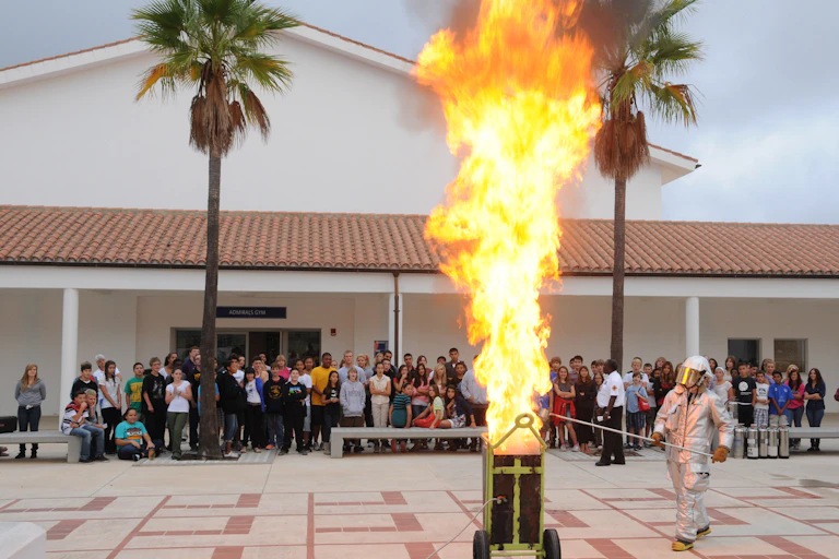 A person in a fire suit standing next to a large flaming pot of oil.
