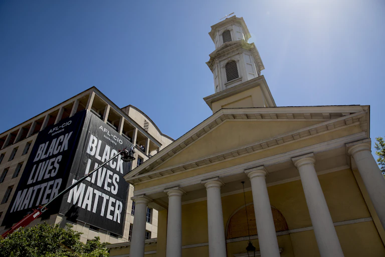 A huge "Black Lives Matter" sign hangs from a building next to a yellow church.