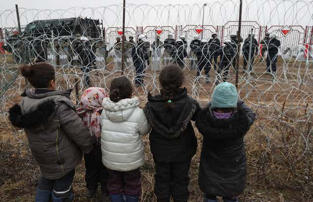 A group of migrant children in warm winter clothes look through barbed wire to a line of uniformed border guards.