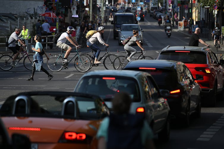 People ride bicycles across a roadway as cars wait.