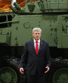 A man in a dark suit, red tie and white shirt with grey hair and glasses stands in front of a light armoured vehicle.
