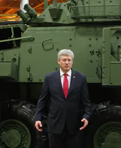 A man in a dark suit, red tie and white shirt with grey hair and glasses stands in front of a light armoured vehicle.