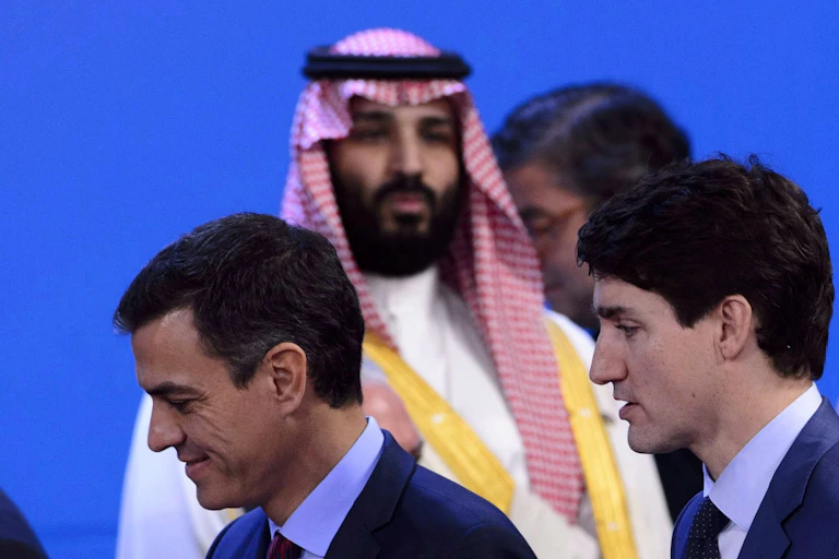 A bearded man in dressed in a Saudi Arabian thobe looks towards two men walking past him in navy suits and ties.