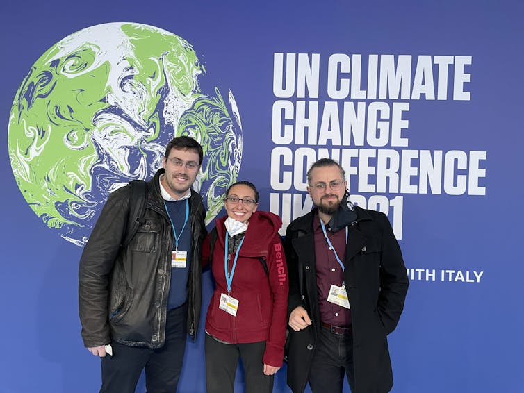 Une femme entourée de deux hommes devant une affiche de la COP26