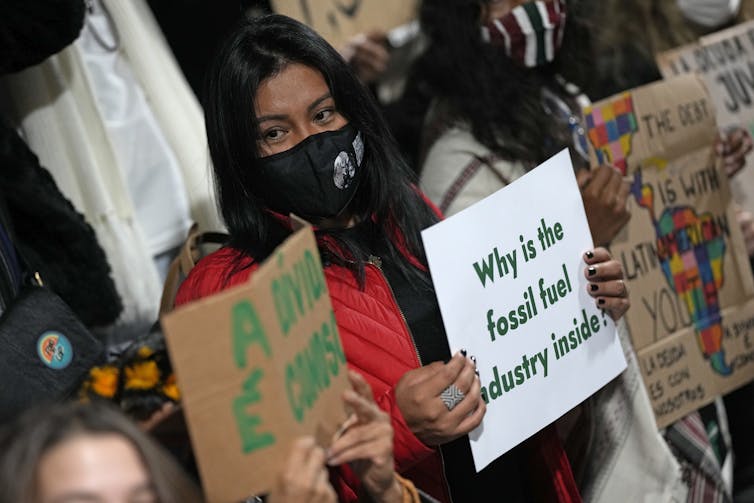 A woman wearing a red jacket holds a sign that reads: Why is the fossil fuel industry inside?