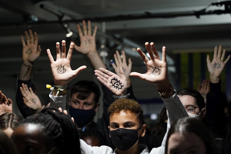 A group of people with their hands in the air. Their palms have 1.5 or an eye drawn on them in black marker.