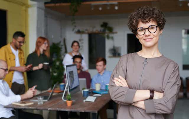 A woman wearing glasses and a taupe shirtsmiles in an office setting with a team of employees behind her.