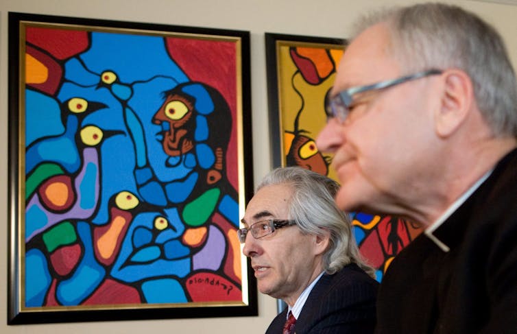 Two men, one Indigenous with longer hair in a suit, and one white man in a clerical priest's collar, sit together.