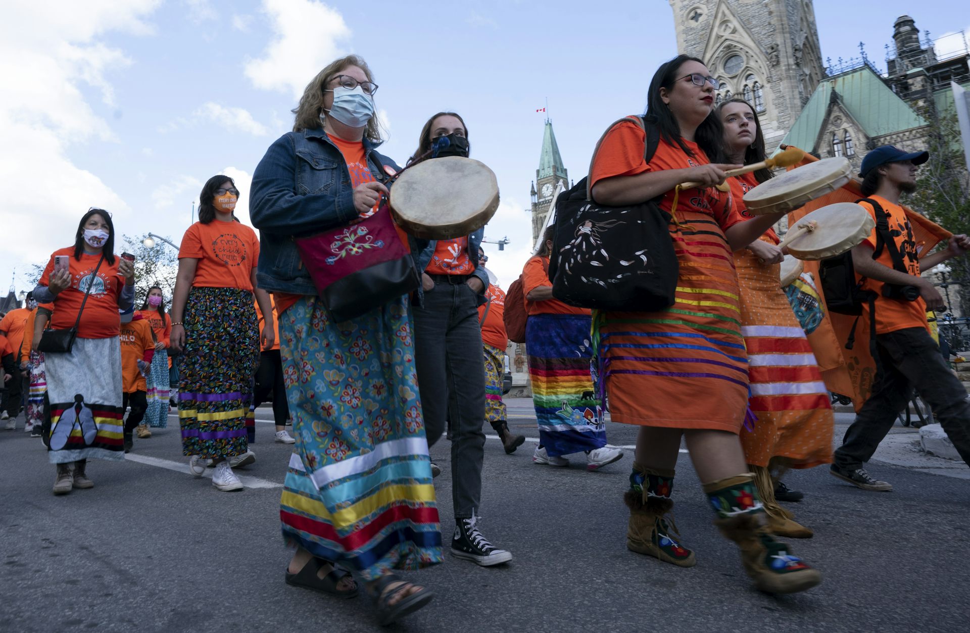 People are seen in orange t-shirts, some wearing ribbon skirts, walking in the street.