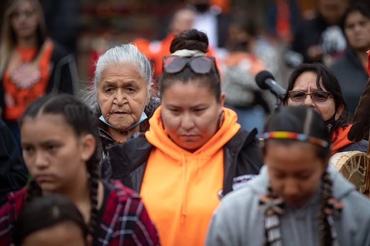 Indigenous women of multiple generations are seen listening in a crowd with sombre faces.
