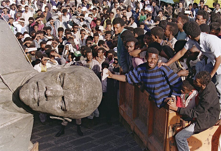 A cheering crowd surrounds a toppled statue