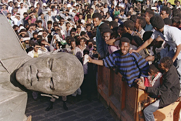 A cheering crowd surrounds a toppled statue