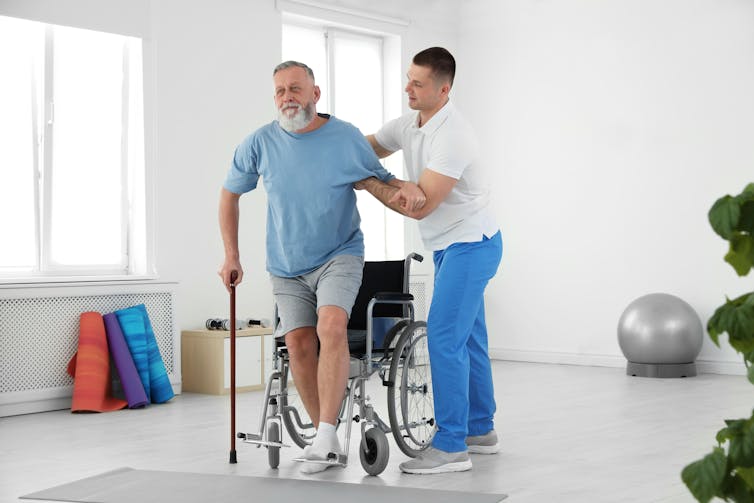 Young male nurse helps an older man stand up from his wheelchair and walk with a cane.
