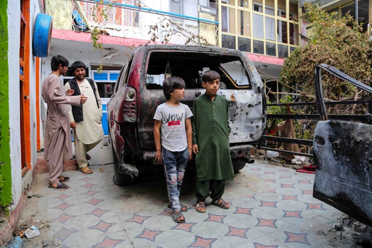 Two boys stand next to a wrecked car with two adult Afghan men in the background. The car was hit by a drone strike in August 2021.