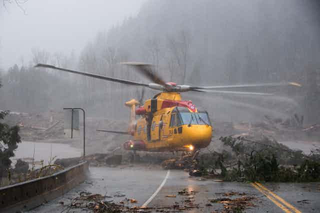 A yellow rescue helicopter lands on a road strewn with branches in heavy rain.