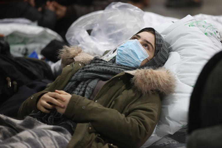 A young woman refugee wearing a face mask and headscarf rests her head on what looks like a large quilt or blanket.