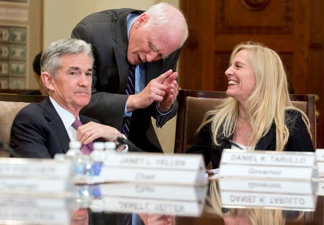 Jerome Powell, seated, looks toward the camera while Lael Brainard, also seated, laughs and looks toward Daniel Tarullo, who is standing and leaning over between them