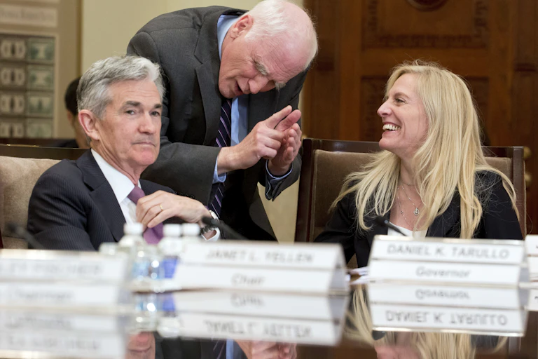 Jerome Powell, seated, looks toward the camera while Lael Brainard, also seated, laughs and looks toward Daniel Tarullo, who is standing and leaning over between them