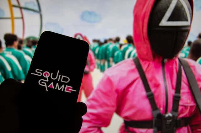 A screen saying SQUID GAME held in front of another screen showing a scene from the show with the players wearing green tracksuits in formation as they are guarded