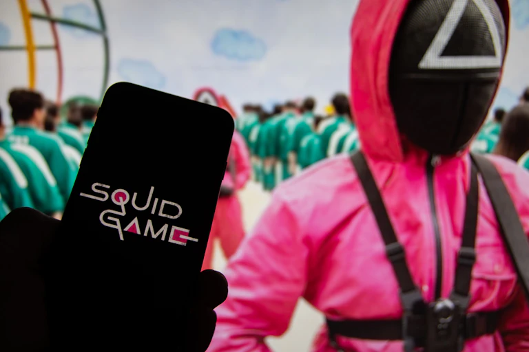 A screen saying SQUID GAME held in front of another screen showing a scene from the show with the players wearing green tracksuits in formation as they are guarded