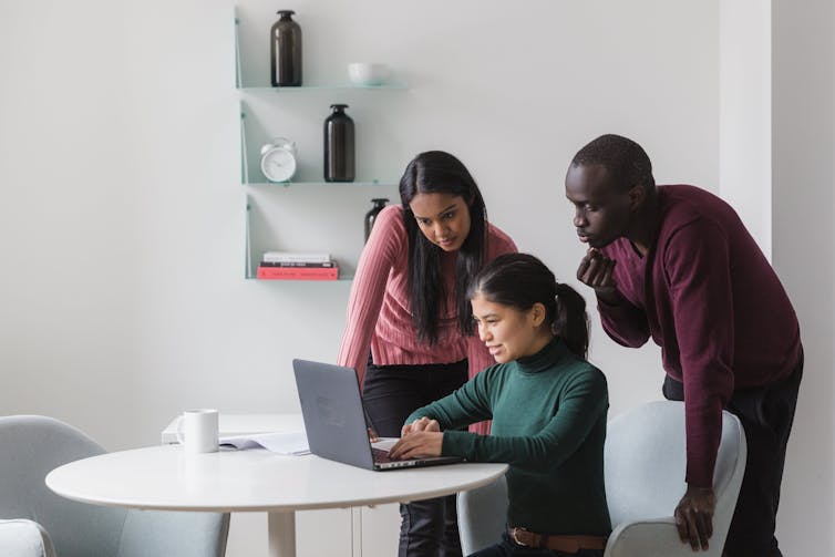 a smiling young woman sits at a table looking at her laptop as two colleagues stand behind her also looking at her screen.