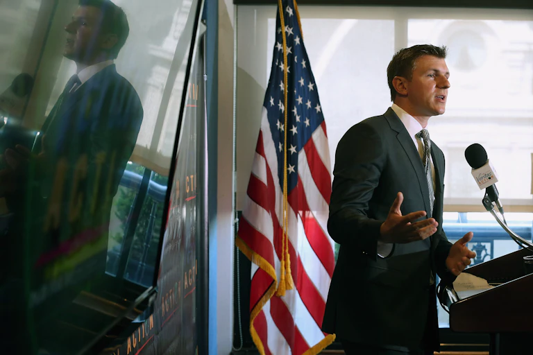 A man in a suit and tie, standing in front of an American flag, speaking into a microphone.