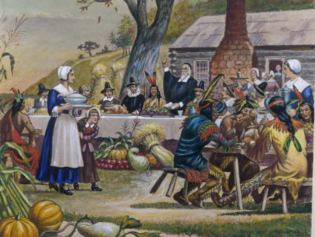 Pilgrims and Native Americans gather at tables to eat.