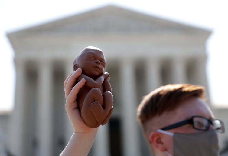 A young man in front of the United States Supreme Court holds up a model of a fetus a little bigger than his hand.