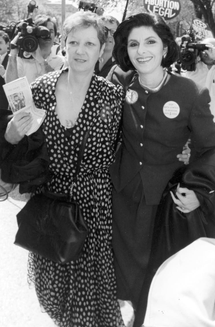 Two women wearing elegant clothes and standing close to each other in front of the Supreme Court.