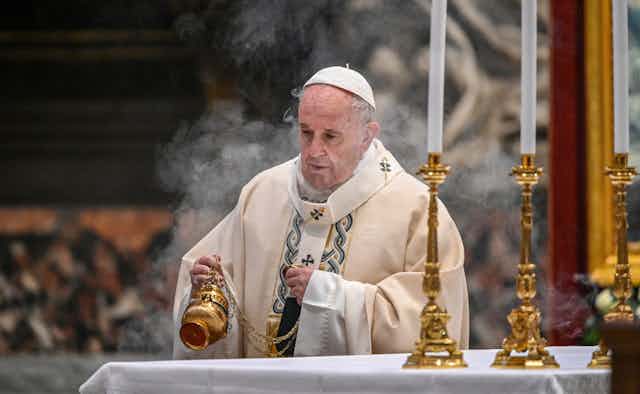 A man in white clerical garments, a liturgical vestment (robe) and skullcap (zucchetto), is seen waving incense over an altar.