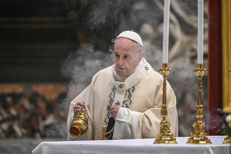 A man in white clerical garments, a liturgical vestment (robe) and skullcap (zucchetto), is seen waving incense over an altar.