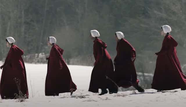 Women in bonnets and red cloaks are seen running across snow.