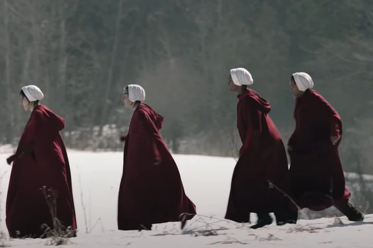 Women in bonnets and red cloaks are seen running across snow.
