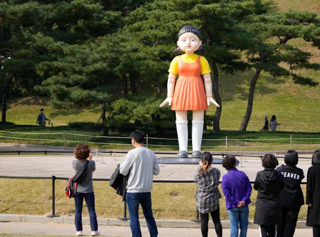 A larger-than-human doll is seen standing in a park and people are looking at it.