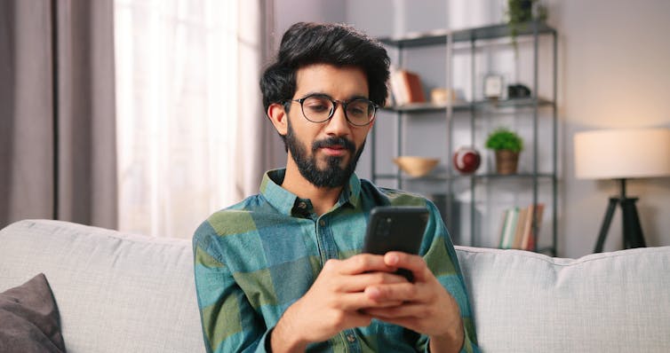 A man sitting on a couch at home typing on his smartphone.