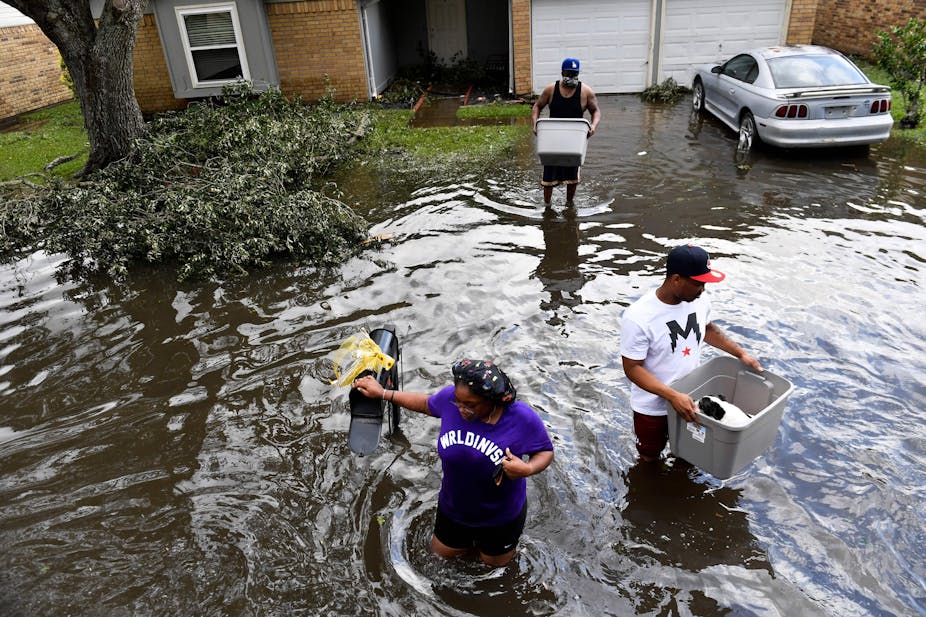 A man carries a dog in a basket and others walk carefully through thigh-high water as they bring belongings out of a flooded home.