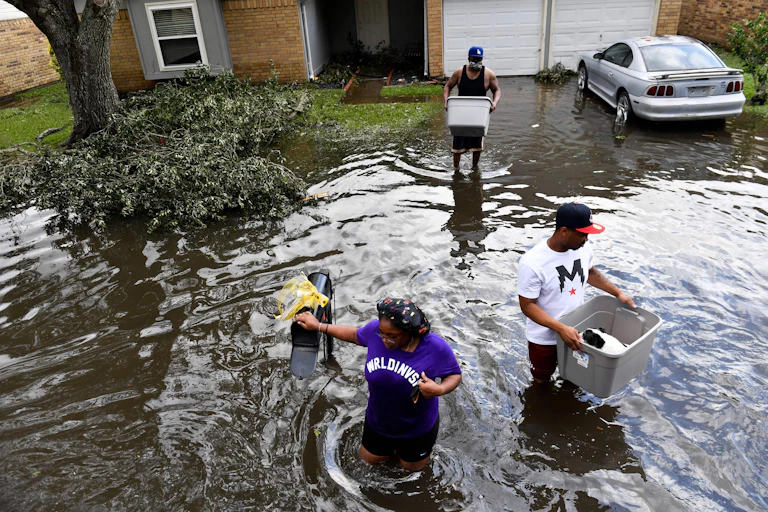 A man carries a dog in a basket and others walk carefully through thigh-high water as they bring belongings out of a flooded home.