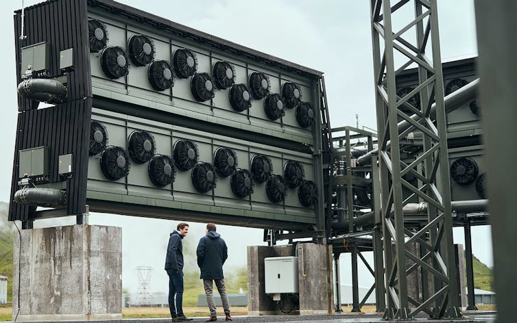 Two men stand beneath a large metal carbon capture structure with fans